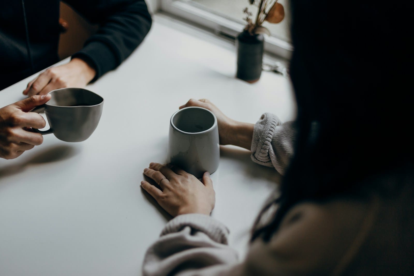 Two people talking across a white table, each holding a grey ceramic mug.