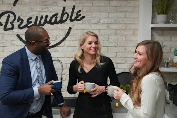 Three colleagues laughing over coffee in an office kitchen.