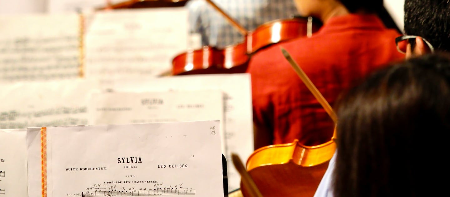 Orchestral players and open sheet music on stands under warm stage light.